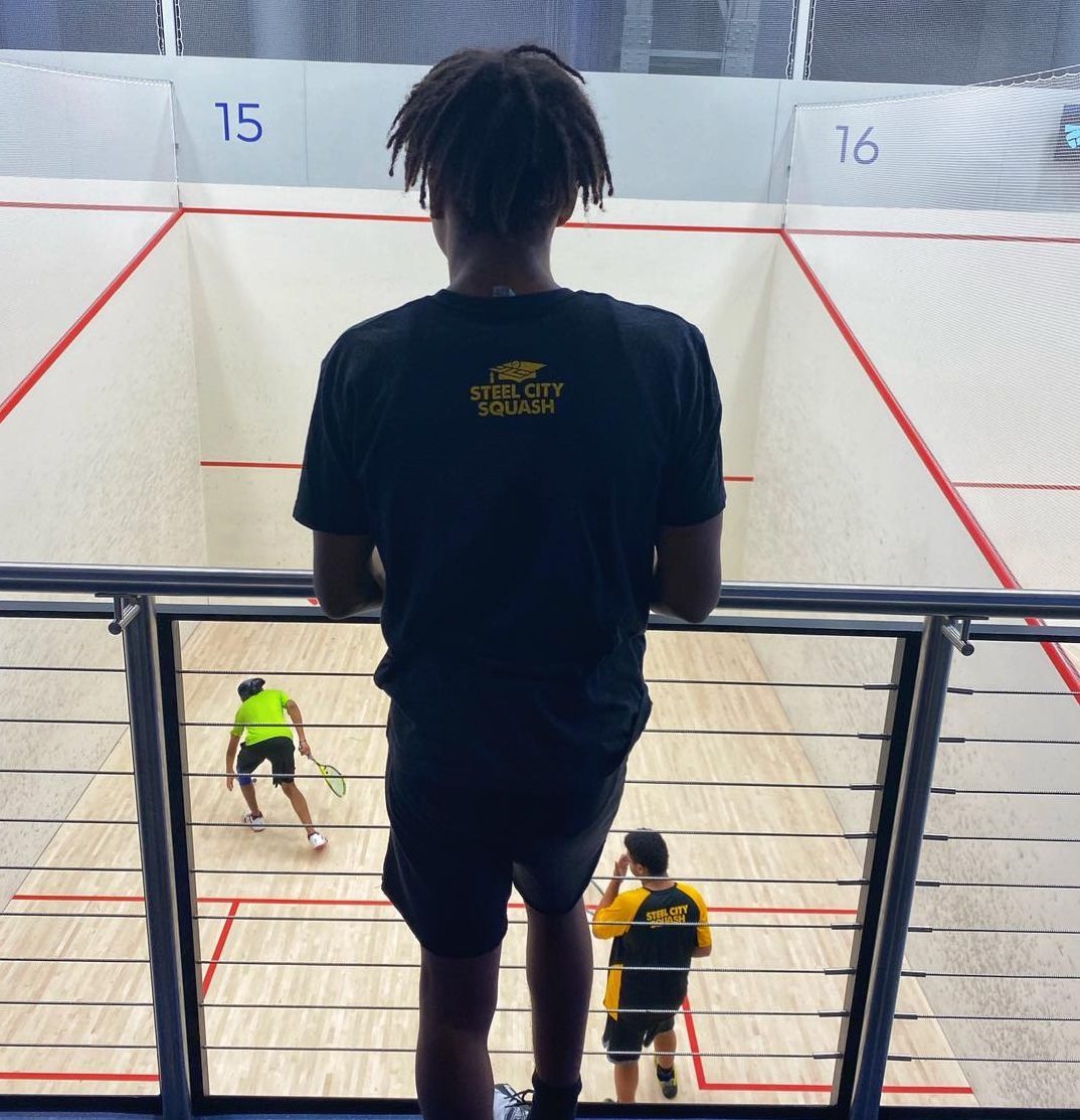 The image shows a young person standing on a balcony overlooking two squash courts where players are actively engaged in a game.