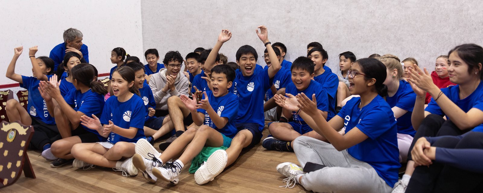 A group of children in blue shirts sit indoors, cheering and clapping enthusiastically. They appear excited and joyful.
