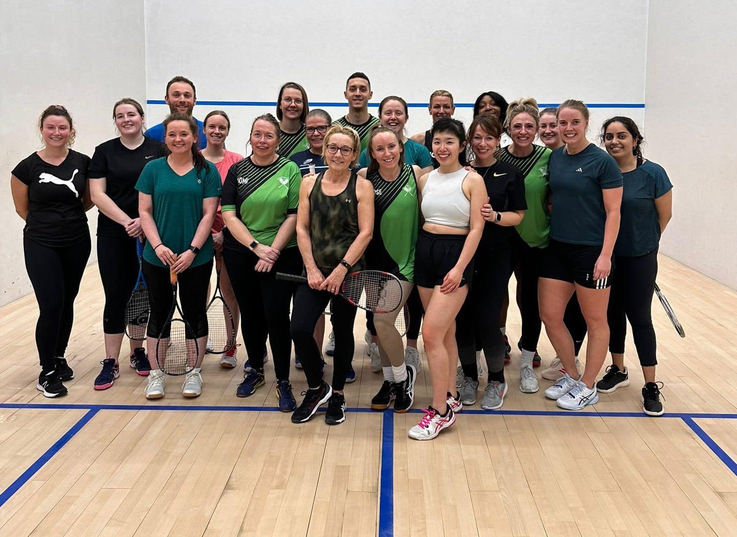 The image shows a large group of squash players posing together on a squash court. The group is diverse, with both men and women, many holding squash racquets, indicating they are either part of a training session or a friendly match.