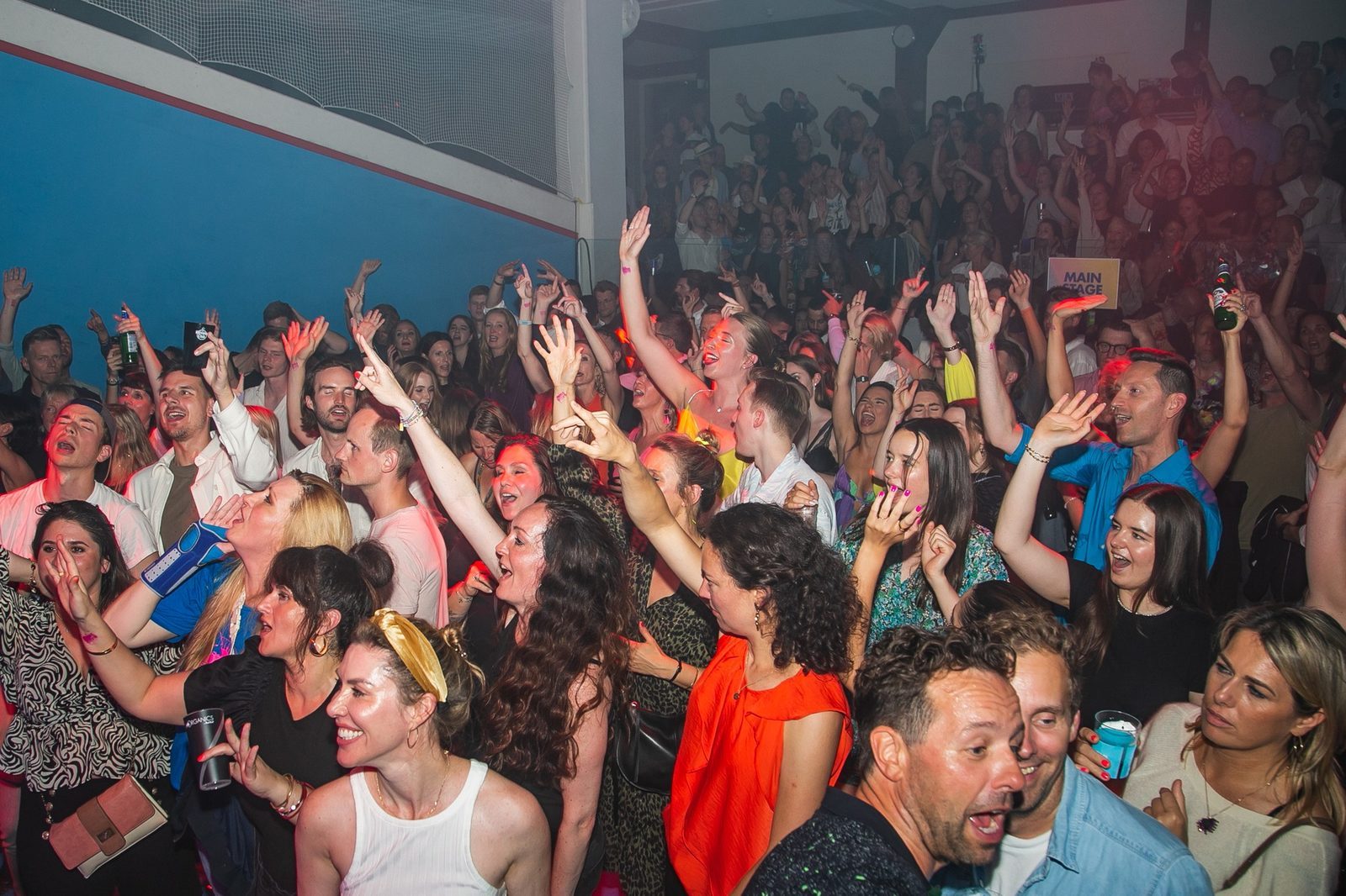 Crowd of people at a vibrant indoor party, cheering with hands raised. Bright outfits, mixed emotions, and a "Main Stage" sign visible.