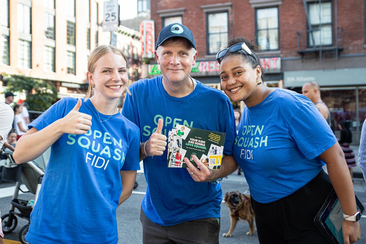 The image shows a group of enthusiastic Open Squash team members engaging in street marketing for the Pearl Street location. Wearing branded t-shirts, they smile and give thumbs-up while handing out promotional materials, creating a positive and energetic vibe to attract potential players and spread the word about the club.