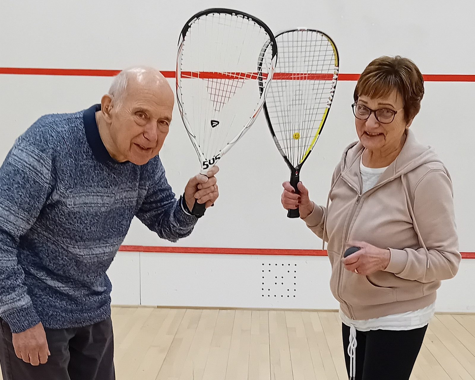 The image shows Christel and Rolf Finkenbrink proudly holding up their Squash 57 rackets inside a squash court. Both are smiling, with Christel to the right dressed in a light hoodie and dark pants, and Rolf to the left in a blue sweater and darker trousers.