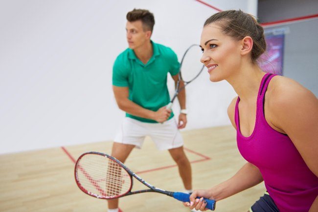 Two people playing squash indoors. Woman in pink tank top, man in green shirt, both holding rackets. Focused expressions, wooden floor.