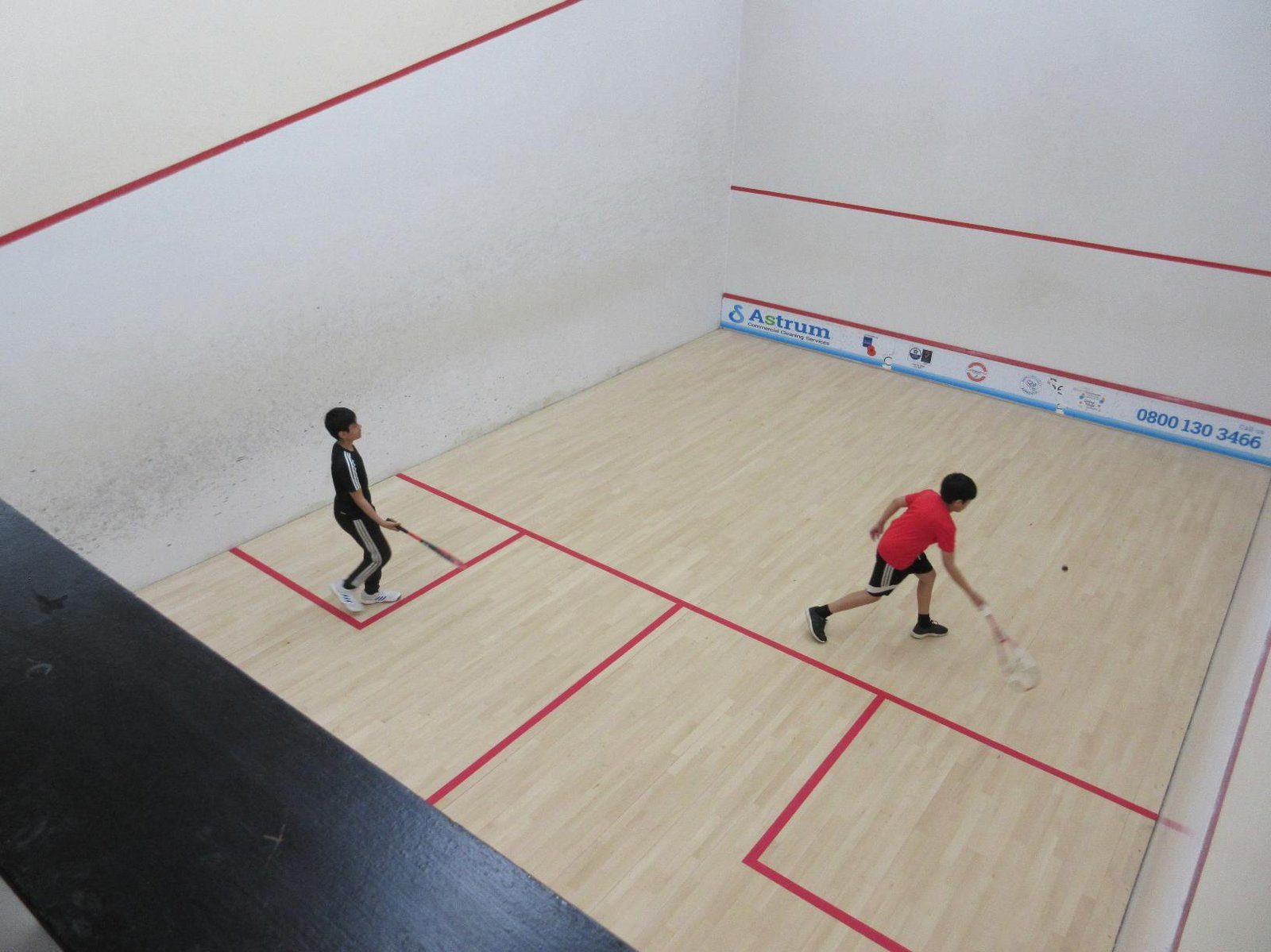 Two boys play squash on a light wood court with red lines. One in a red shirt lunges to hit the ball, while the other watches.