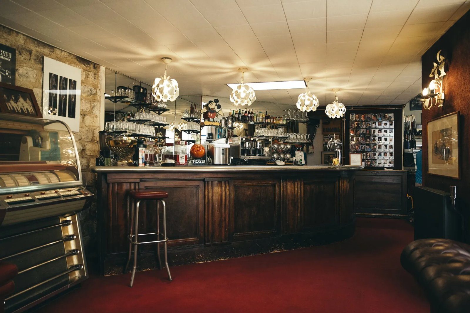 Cosybar with a wooden counter, red carpet, retro jukebox, and hanging lights. Shelves filled with glasses and bottles behind. Cozy ambiance.