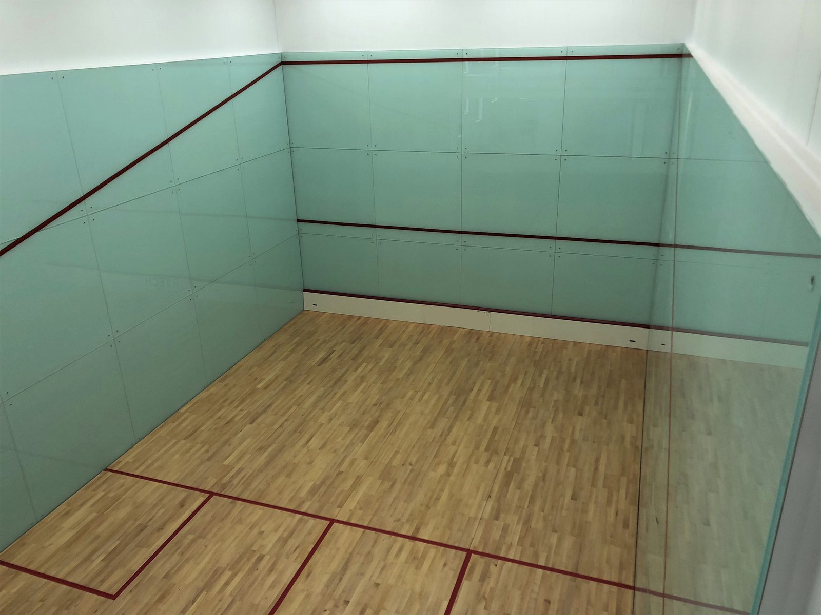 squash court with light blue glass walls and wooden floor, marked with red lines. The space is bright and clean, creating a calm atmosphere.