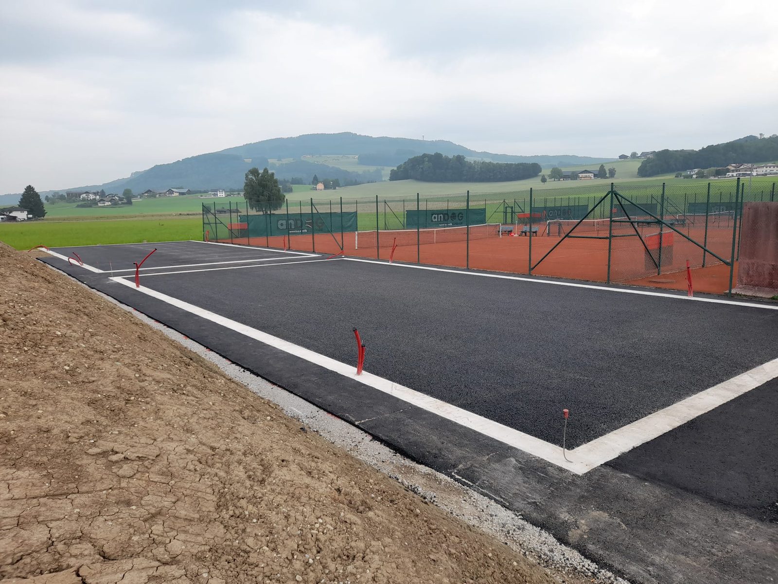Freshly paved outdoor padel court base with white edge markings, located beside a red clay tennis court in a rural area with hills and farmland.