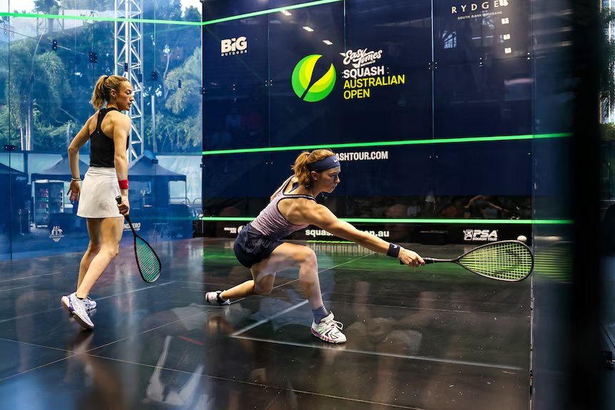 Two women playing squash in a glass court. One lunges to hit the ball. Squash Australian Open logo visible. Intense action and focus.