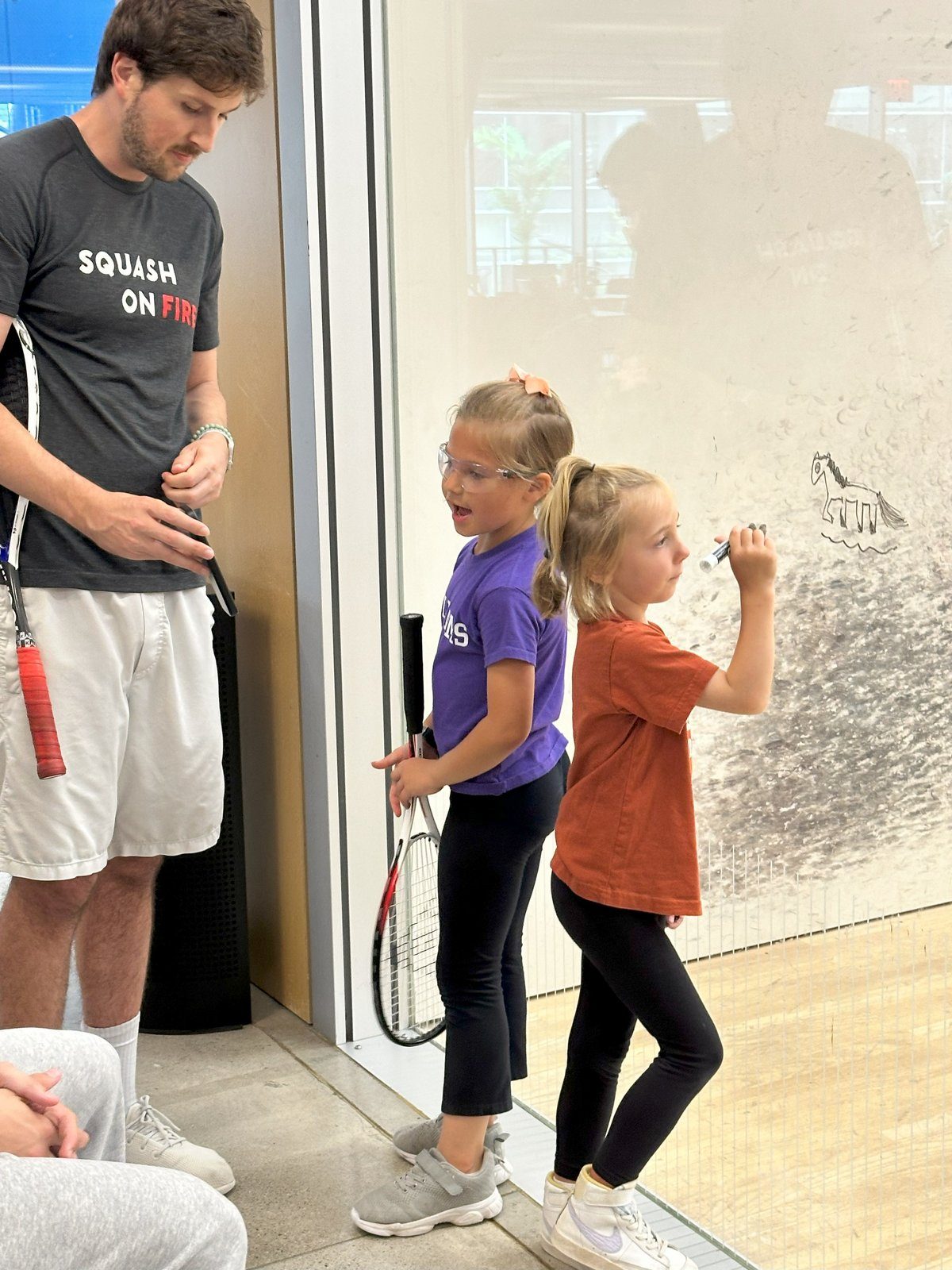 Man with a "Squash On Fire" shirt talks to two girls with rackets. One draws on a glass wall. Indoor sports setting, casual mood.