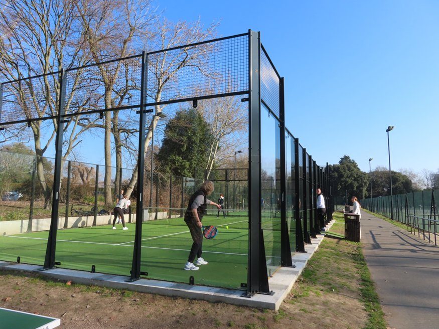 Outdoor, glass-walled padel courts with a doubles game in progress on green turf; additional players and two onlookers stand along a walkway under a clear, sunny sky.