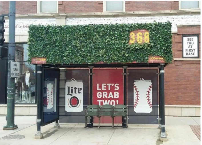 Bus stop with green foliage roof, red and white baseball-themed ads. Text reads "LET'S GRAB TWO!" and "SEE YOU AT FIRST BASE."