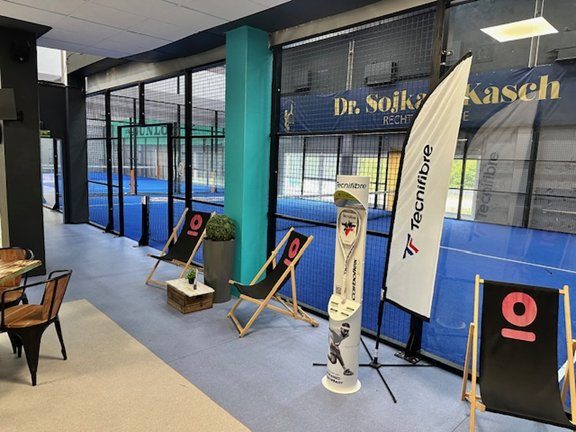 Indoor padel courts with lounge chairs, banners, and Tecnifibre display in a modern, welcoming viewing area.