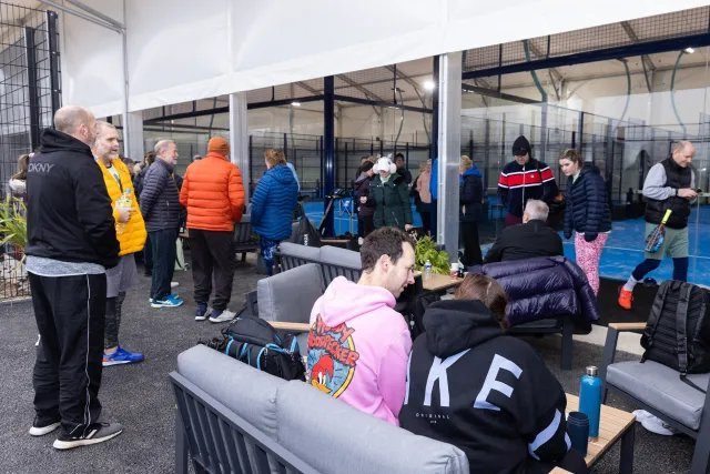 People in colorful jackets chat and relax on couches in a bright indoor sports facility. Background shows courts and a casual atmosphere.
