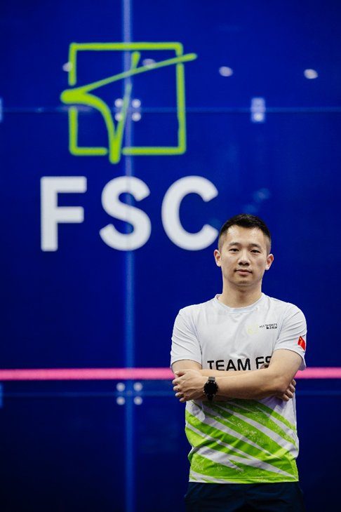 Jack Wang wearing a "TEAM FSC" shirt stands confidently with arms crossed in front of a glass squash court wall displaying the Tengfei Squash Club (FSC) logo.