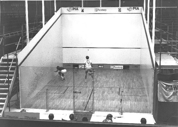 Two players in a squash court; one lunges for the ball. The court is enclosed with glass walls. Visible logos include PIA and Perstorp.