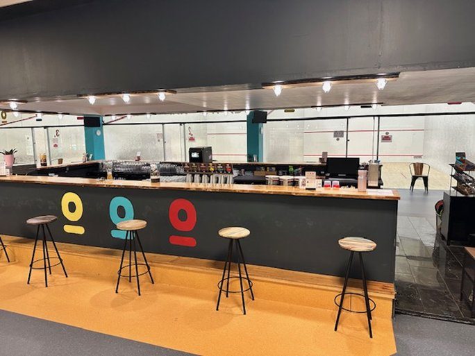 Stylish bar area with colorful icons and high stools, overlooking squash courts in a modern indoor sports facility.