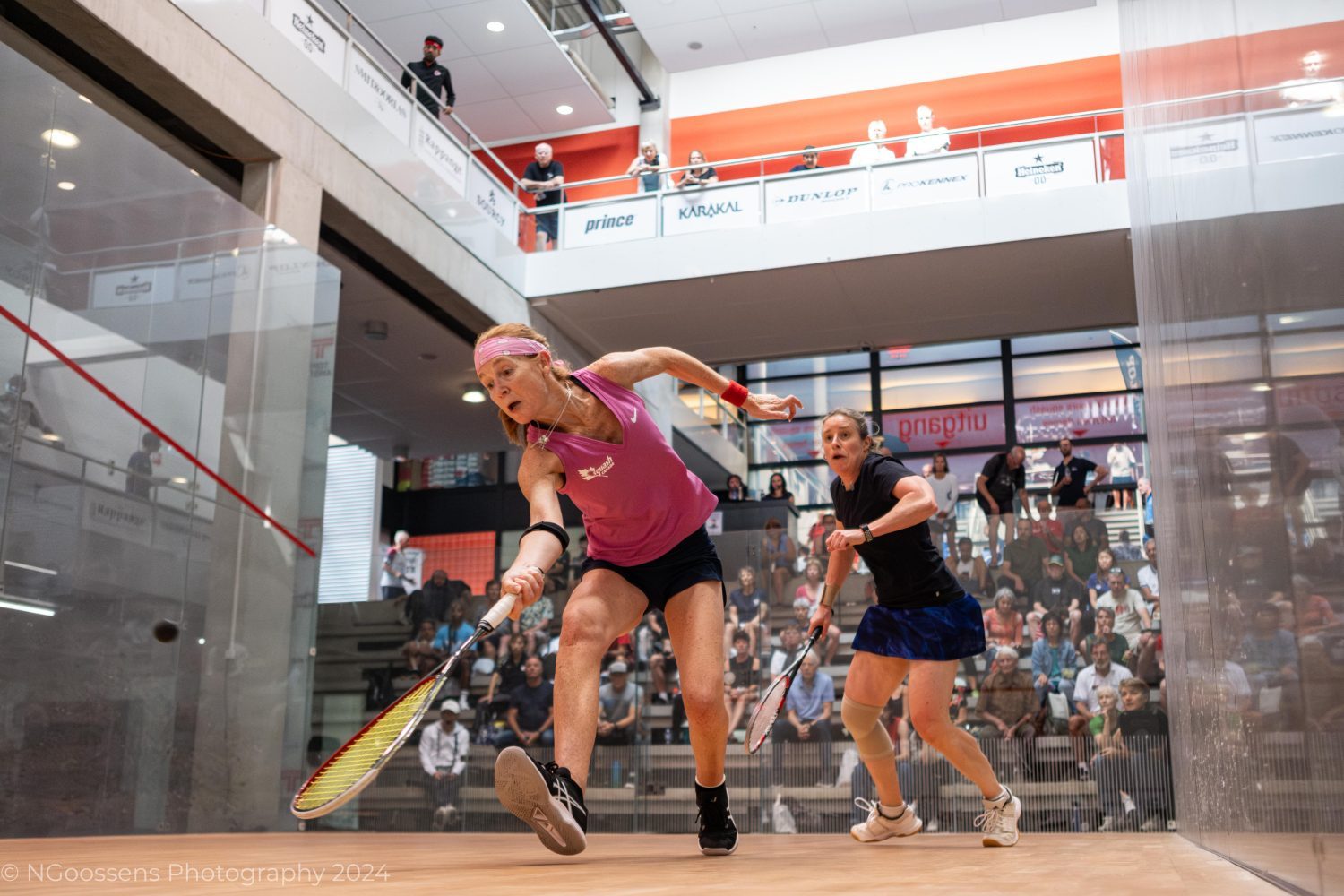 The image captures an intense moment of action on the squash court. Two players are fully engaged in a competitive match, with the player in the foreground lunging forward to reach the ball with her racquet.