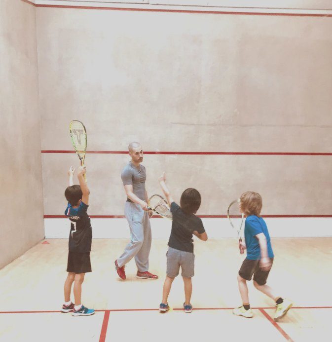 A coach and three kids with rackets in a squash court. The coach instructs while kids point and listen, wearing colorful sportswear.