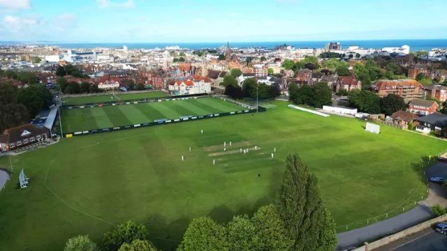 Aerial view of a cricket match on a green field. Players are scattered across the pitch. City buildings and trees form the background.