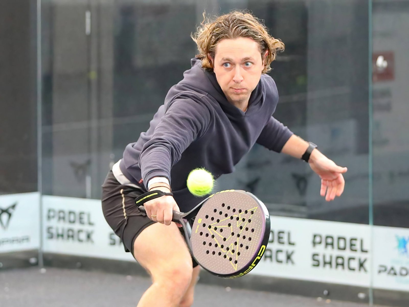 Man playing padel in action, focused expression. Wearing hoodie, swinging racket at ball.
