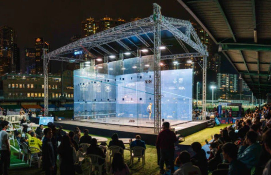 Outdoor glass court, large crowd watching. City skyline illuminated in the background.