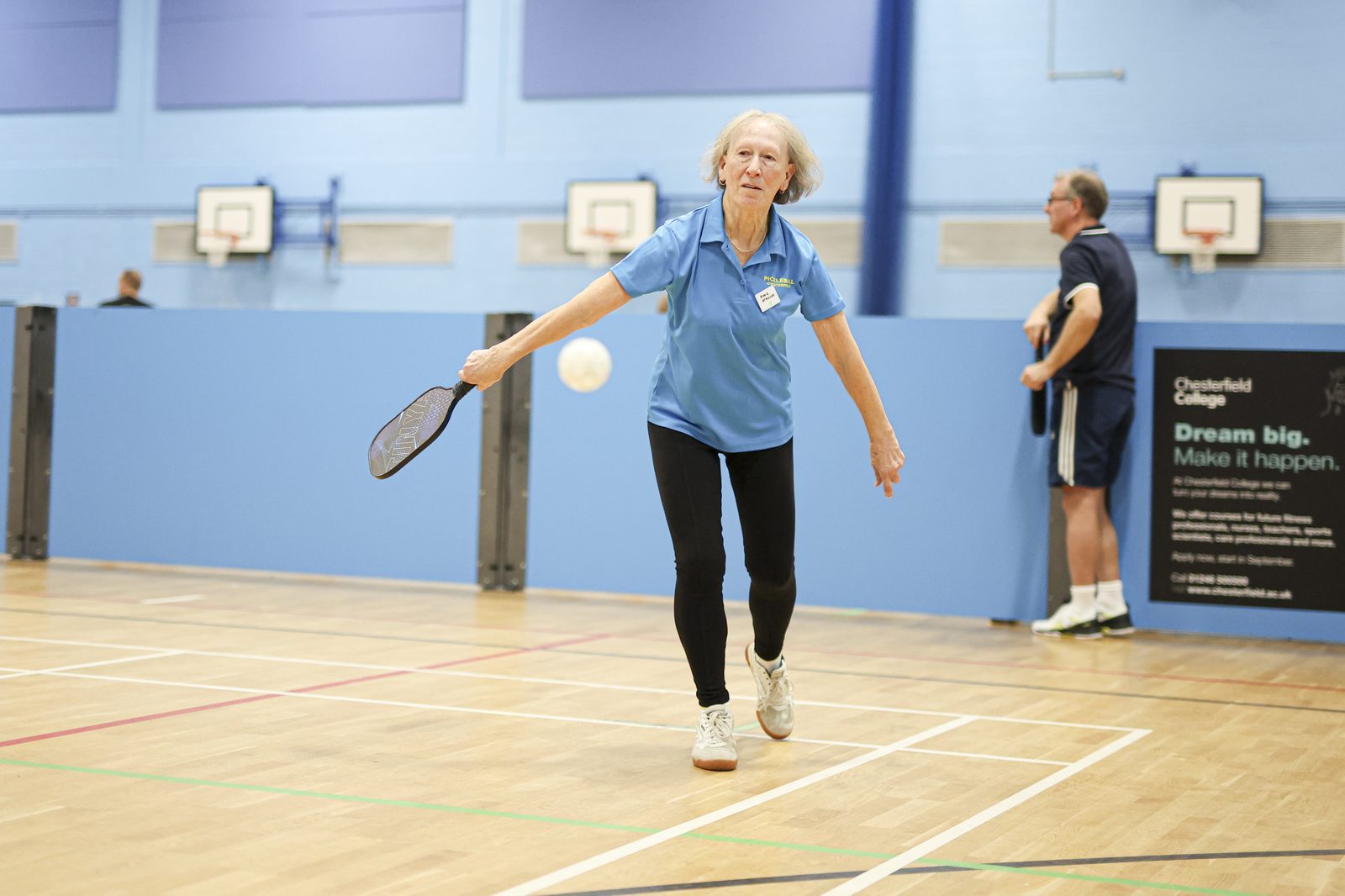 Elderly woman playing pickleball in a gym, wearing a blue shirt. Background shows blue walls, a basketball hoop, and motivational text.