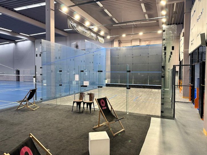 Modern glass squash court with seating area and lounge chairs, adjacent to badminton courts in a bright, spacious indoor sports hall.