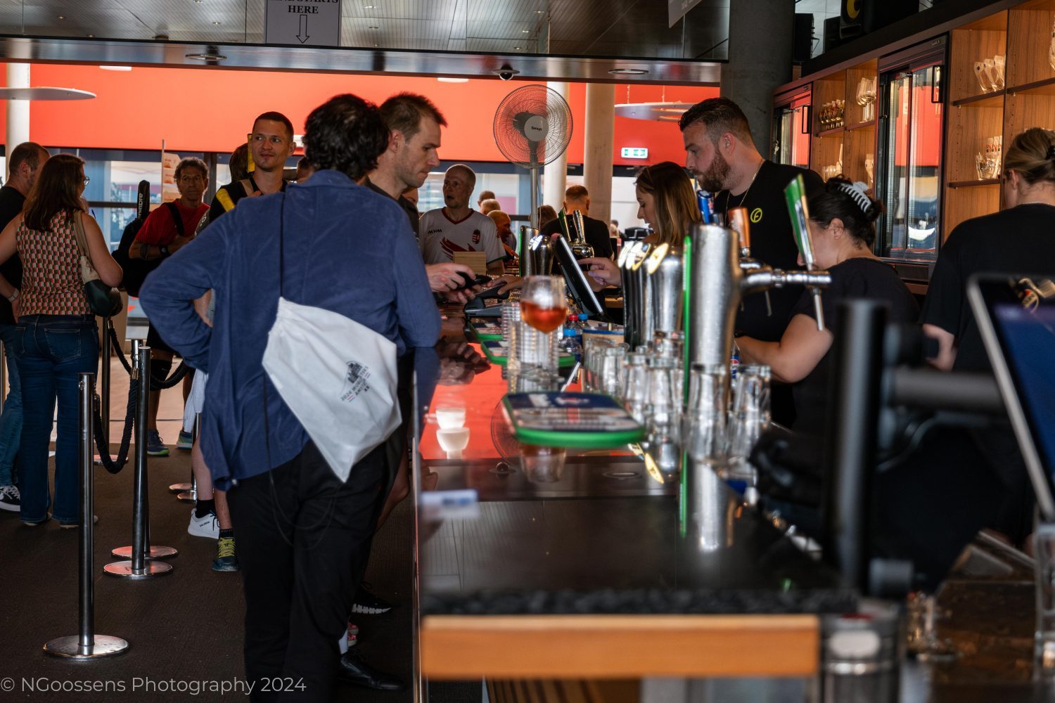 The image shows the bustling bar at the Frans Otten Stadion, a central social hub for members and visitors. Several people are gathered around the bar, ordering drinks and engaging with the staff.