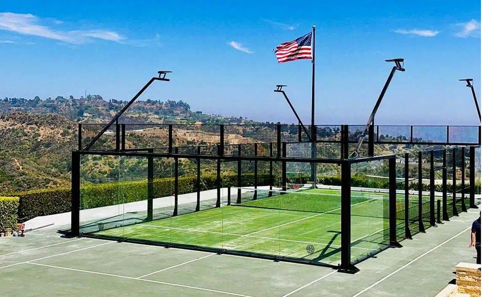 Outdoor panoramic padel court with green artificial turf and minimal corner posts, offering unobstructed views through large glass panels. Located on an elevated terrace with scenic hillside views, the court features integrated LED lighting and an American flag in the background under a clear blue sky.