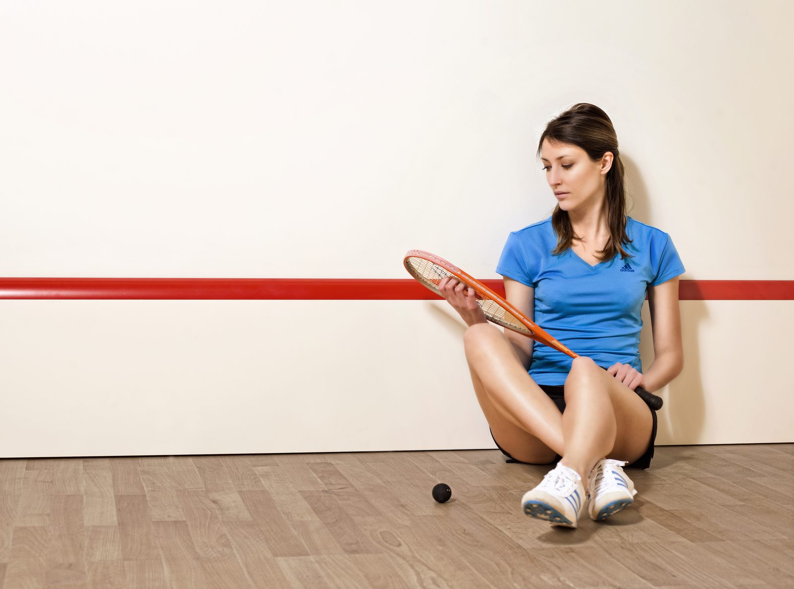 Woman in blue shirt, sitting on a squash court floor, holding a racket. A black squash ball lies nearby. Neutral expression.