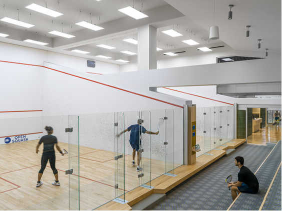 Indoor squash facility with courts at Open Squash, players in action and one seated viewer observing the match.