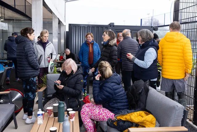 Group of people in winter jackets socializing and using phones on a patio. Coffee cups on table, casual setting with a mix of colors.