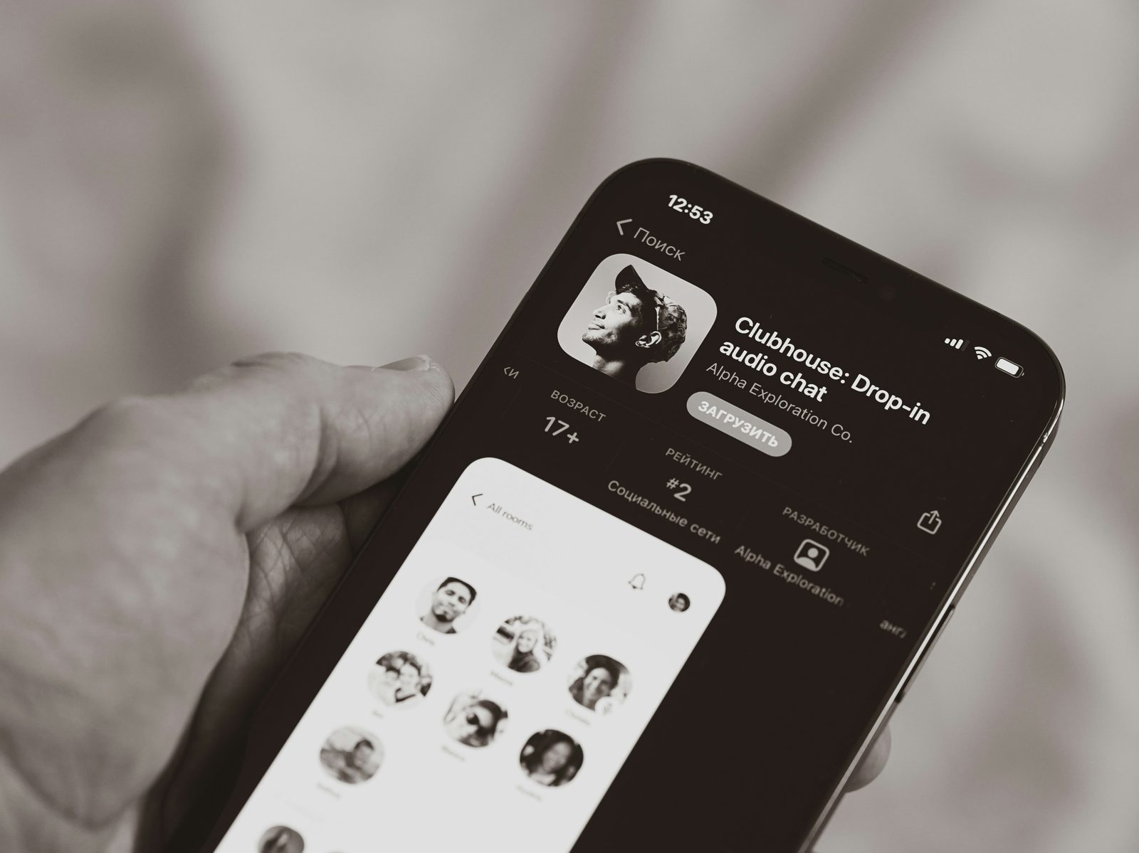 Close-up of a hand holding a smartphone displaying the Clubhouse app page and interface, shown in black and white.