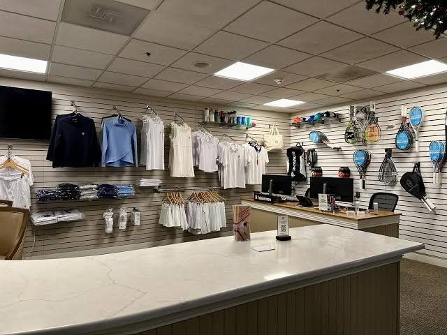 A well-organized pro shop inside a racket sports facility. The shop features a reception counter in the foreground, neatly displayed tennis and squash apparel on the left wall, accessories and grips above the counter, and a variety of rackets hanging on the right wall.