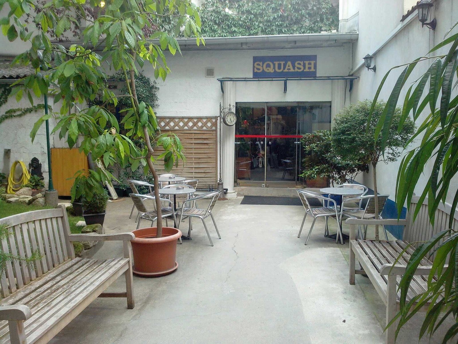 Outdoor patio with metal tables and wooden benches, surrounded by potted plants. "SQUASH" sign above a glass door. Peaceful mood.