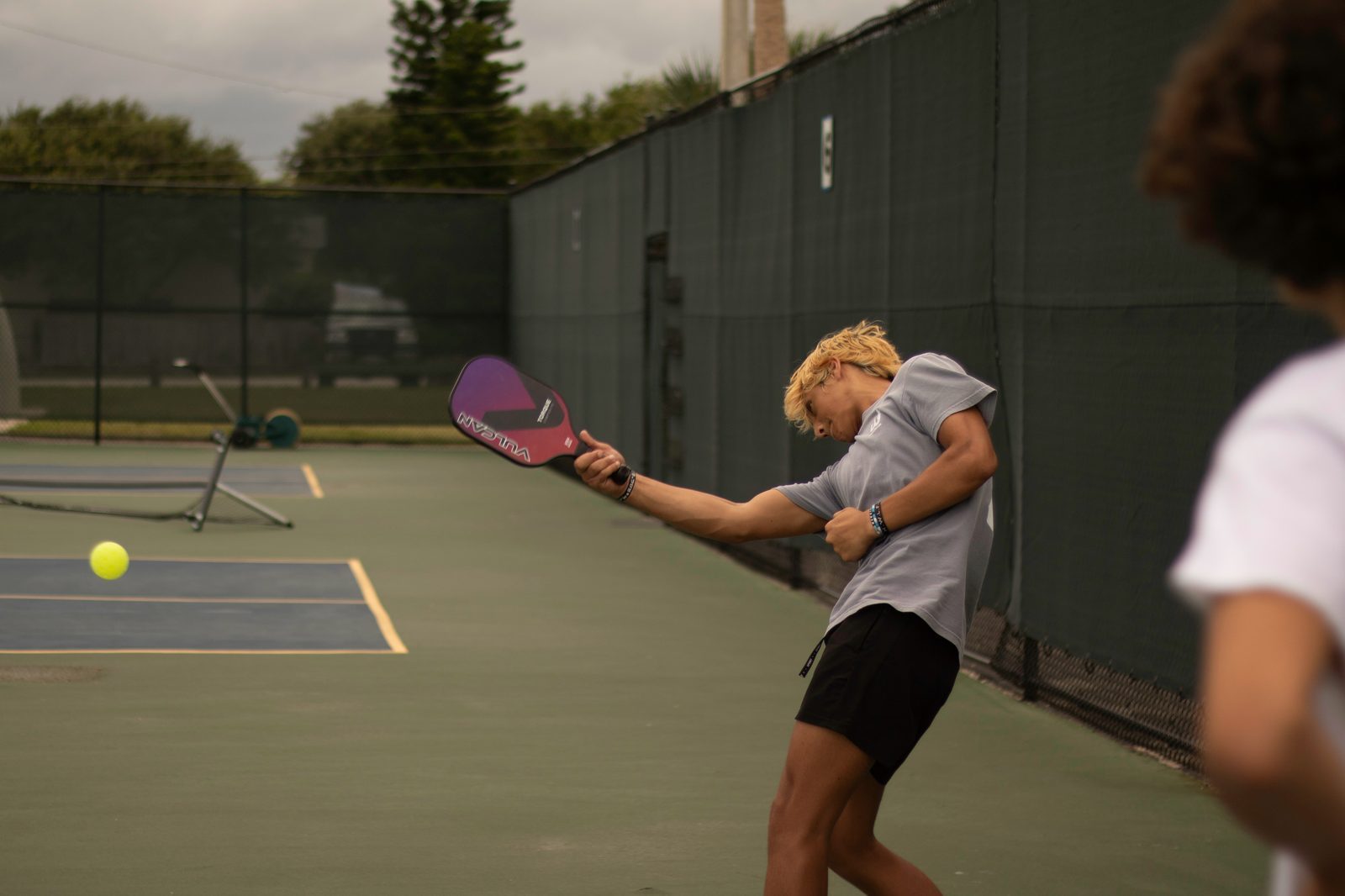 A young person with blonde hair in athletic attire plays pickleball on an outdoor court. They are mid-swing, hitting a ball with a purple paddle. Another player, slightly blurred, is visible in the foreground. The scene is set on a fenced-in court with trees and buildings in the background under a cloudy sky.