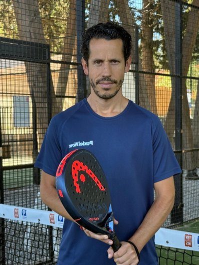 A man standing on a padel court holding a black and red padel racket. He is wearing a dark blue sports shirt and looking toward the camera, with a net and trees visible in the background.