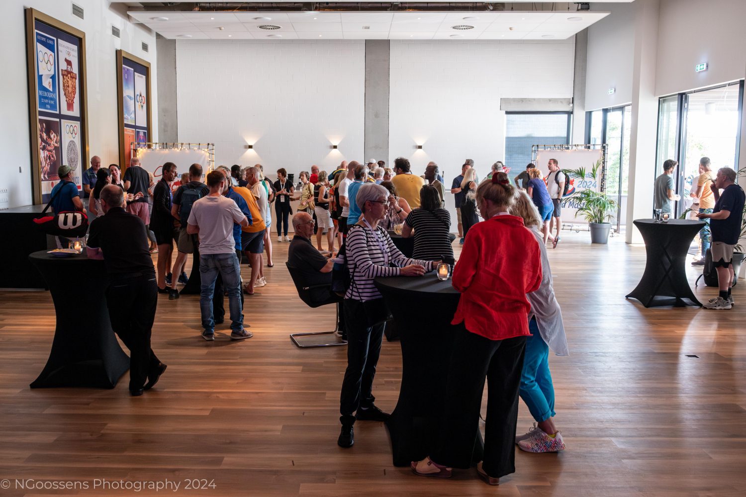 The image captures a lively event during the World Masters 2024. Participants and spectators are gathered in a spacious, well-lit room with wooden flooring, engaging in conversations and networking.