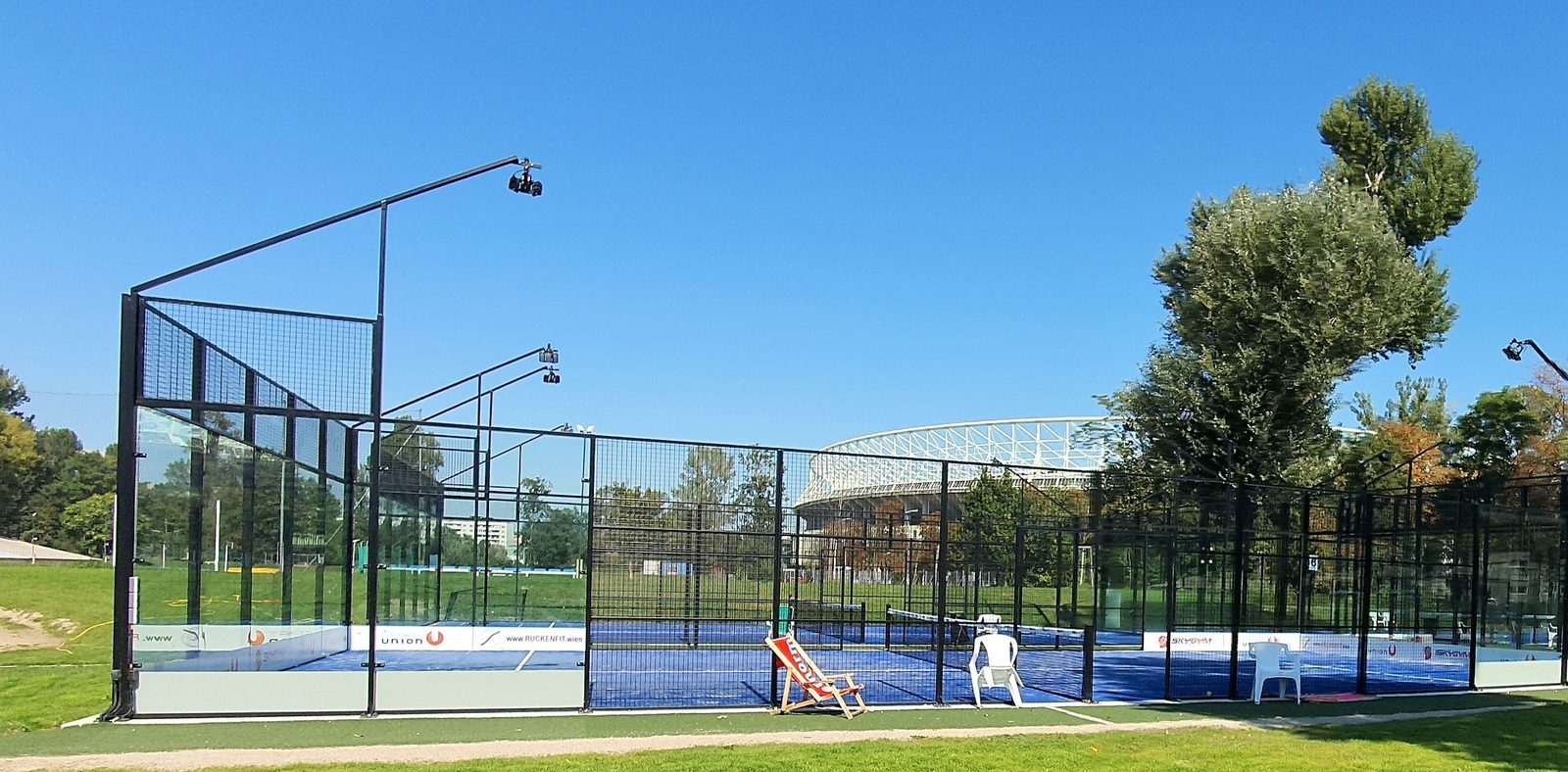 Outdoor padel court in club classic design featuring steel support posts and blue artificial turf, surrounded by metal fencing and tall LED floodlights. The court is set in a green park area under a clear blue sky, with trees and a stadium visible in the background.