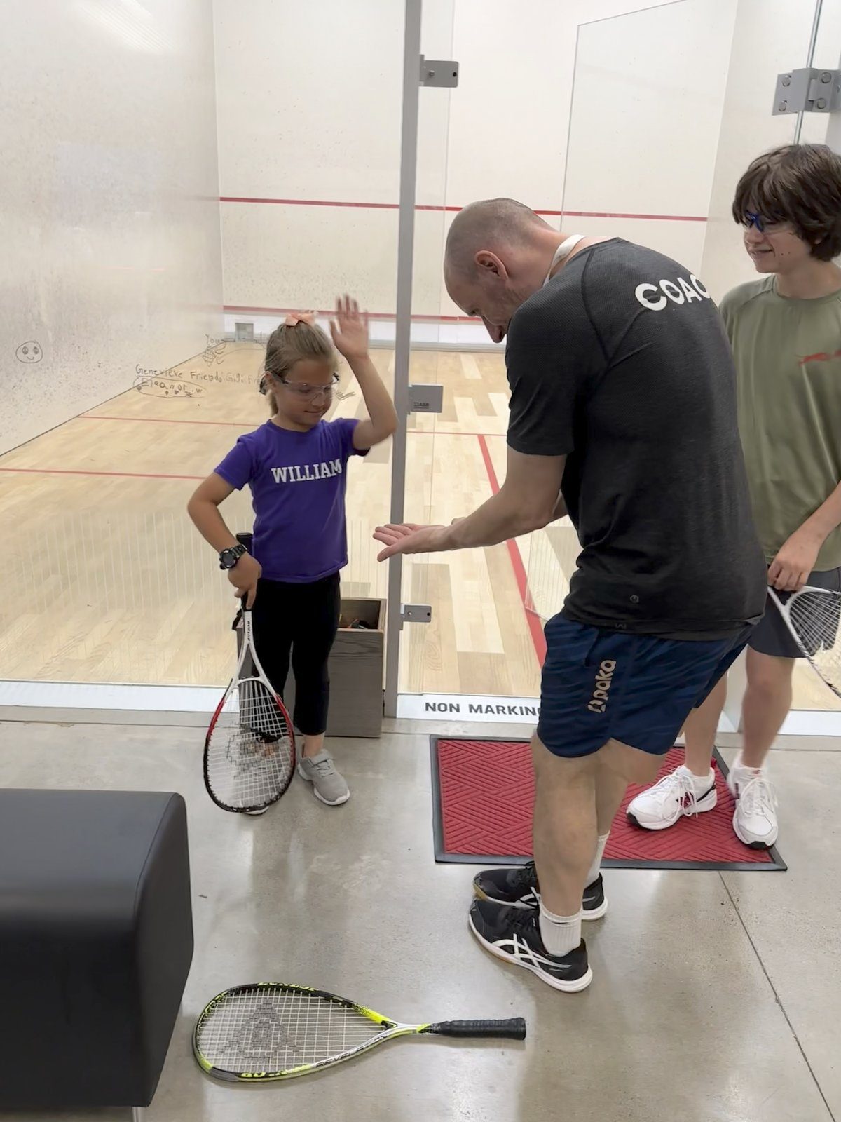 Coach gives feedback to a young junior in goggles on a squash court while another camper waits—small-group instruction at a summer camp.